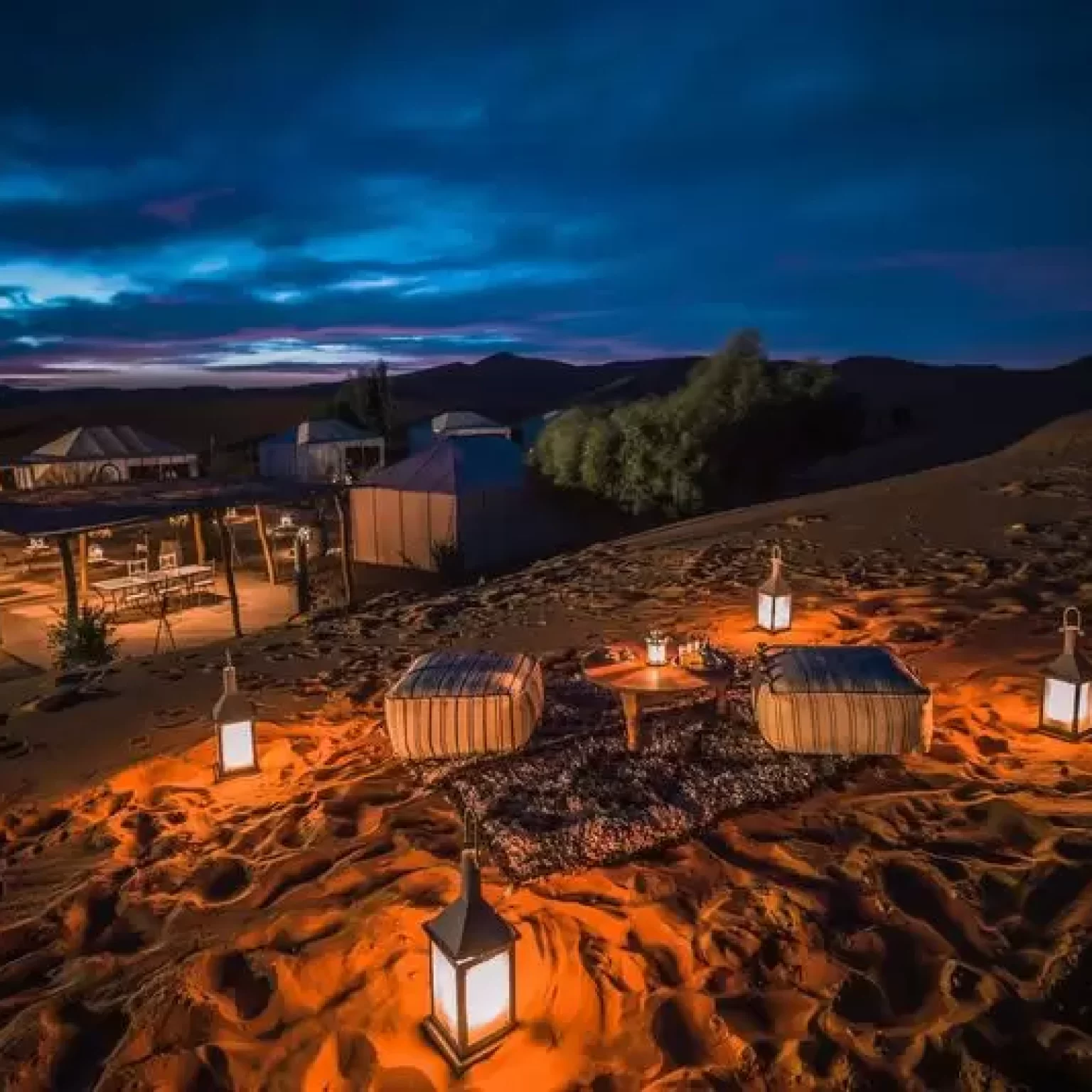 1. A serene desert camp featuring glowing lanterns and a rustic table set against a vast sandy landscape while your 12-day Morocco luxury Tour From Marrakech