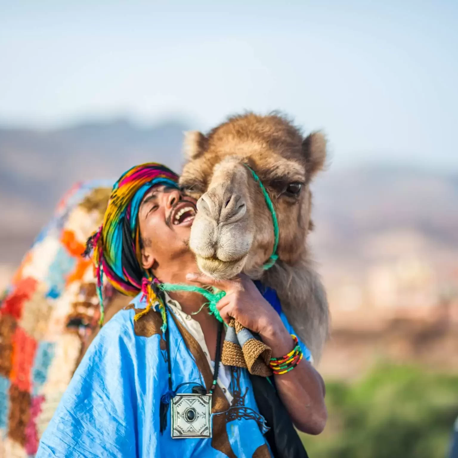 A man in traditional attire smiles beside a camel, showcasing a moment of cultural connection and joy in the desert landscape:Camel rides in Erg Chebbi