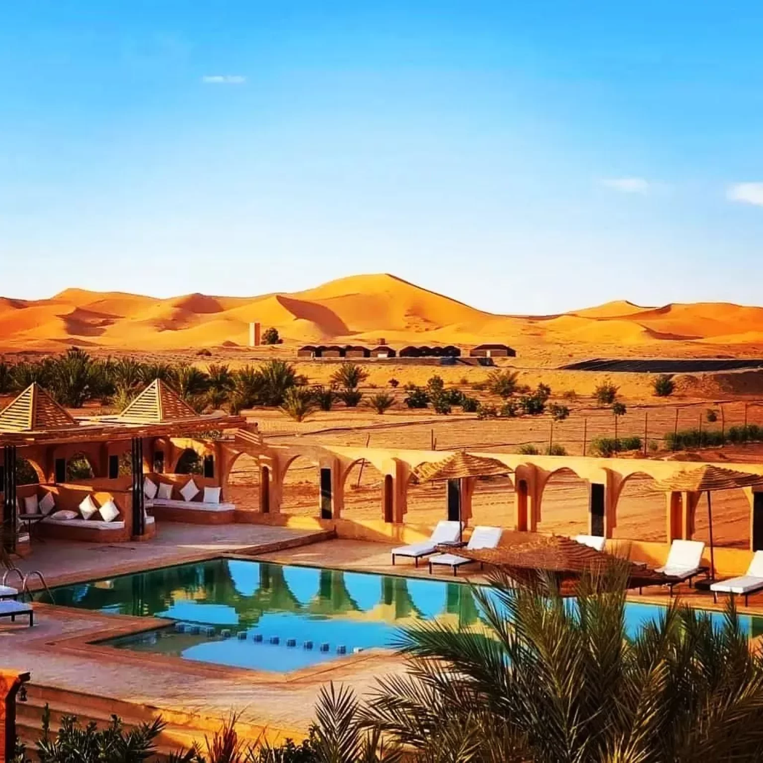 1. A serene desert resort featuring a sparkling pool surrounded by sun loungers under a clear blue sky while your 12-day Morocco luxury Tour From Marrakech