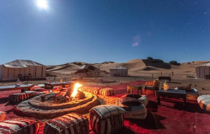  A serene fire pit in the desert of Merzouga, illuminated by a bright full moon in the night sky.