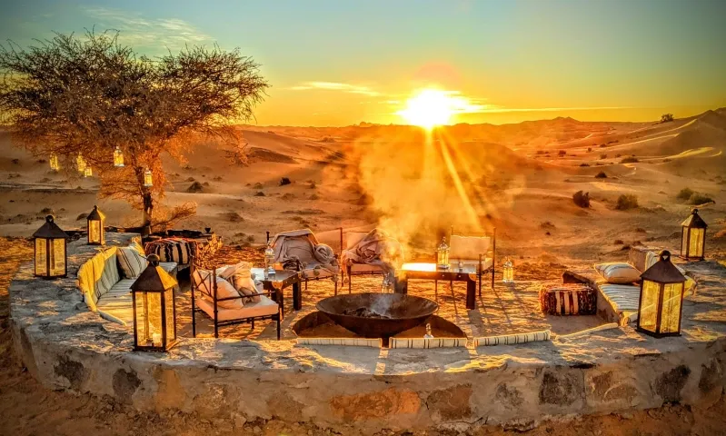 A tranquil sunset in the Desert Camps in Merzouga, highlighting a fire pit and chairs, perfect for evening gatherings.