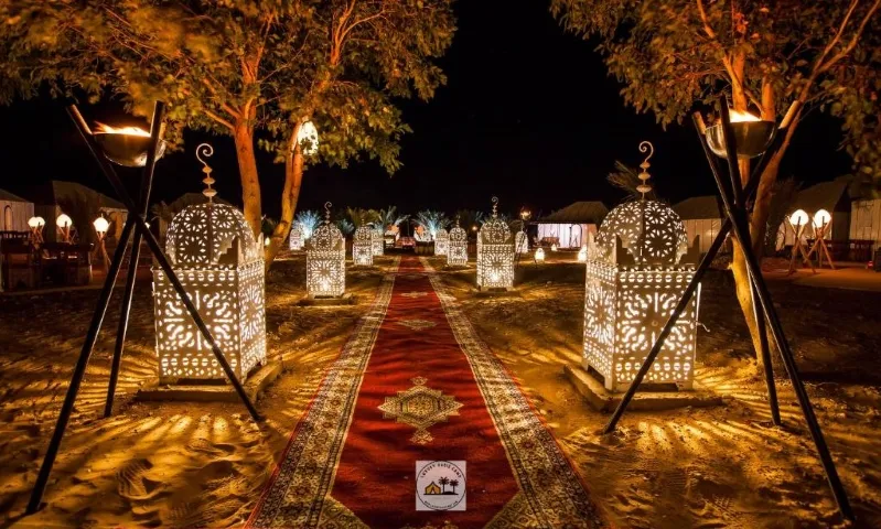 A serene, white carpeted path illuminated by lights and candles, leading through the Desert Camps in Merzouga.