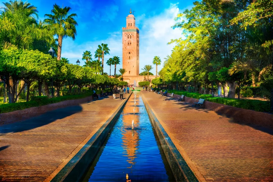 A fountain surrounded by greenery in a park, with a prominent clock tower visible of the Koutoubia Mosque in the background.